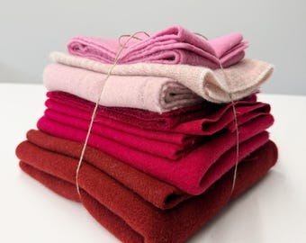 May include: A stack of folded felt fabric pieces in a gradient of colors ranging from light pink to deep red. The fabric layers are secured together with a thin, light-colored string wrapped around the center of the entire textile bundle.