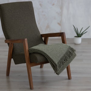Puede incluir: Un sill&oacute;n verde con brazos y patas de madera, una manta de lana verde est&aacute; colocada sobre el sill&oacute;n.