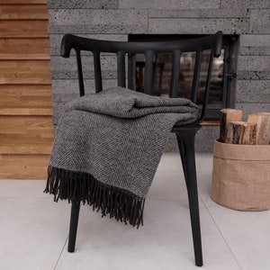 Può includere: Una coperta in lana a spina di pesce grigia e nera con frange drappeggiata su una sedia in legno nero. La sedia è davanti a un camino con un rivestimento in pietra.