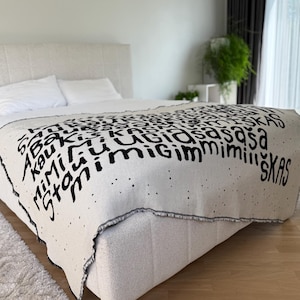 May include: A white throw blanket with black text that reads "skas" and other words. The blanket is on a white bed with white pillows.