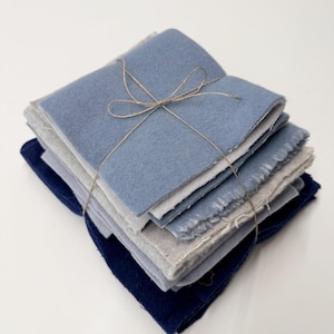 Op de afbeelding: Een stapel vierkante stoffen lappen in tinten blauw, grijs en marineblauw, samengebonden met een dun bruin touwtje in een strik. De stoffen hebben zichtbare, rafelige randen en een zachte, viltachtige textuur, gerangschikt in een nette stapel.