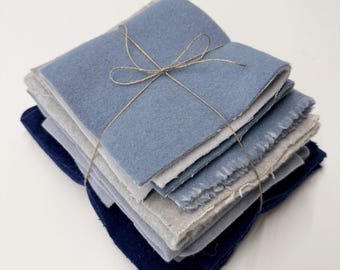 May include: A stack of square fabric swatches in shades of blue, gray, and navy, tied together with a thin brown twine bow. The fabric pieces have visible raw, frayed edges and a soft, felt-like texture, arranged in a neat, layered pile.