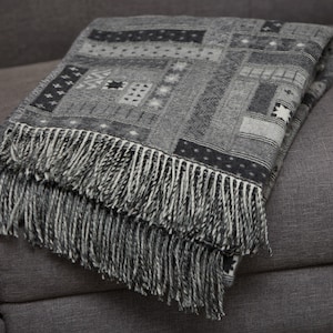 May include: A grey and white patterned throw blanket with fringe on the edges. The blanket is folded and draped over a grey sofa.