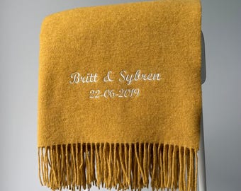 以下が含まれることがあります： Britt & Sybrenという文字と22-06-2019という日付が白く刺繍された、マスタードイエローのブランケット。ブランケットは質感のある表面で、下端にはフリンジが付いており、白いフレームに掛けられています。