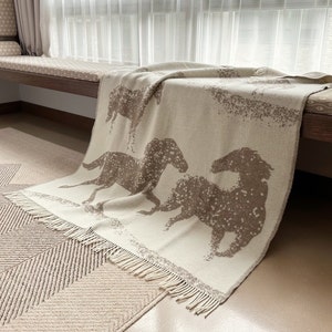 May include: A cream-colored throw blanket with a brown horse design. The blanket has a fringe border and is draped over a wooden bench.