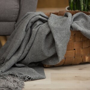 Puede incluir: Una manta de lana gris con flecos est&aacute; colocada sobre una cesta tejida. La cesta est&aacute; hecha de materiales naturales y tiene un color marr&oacute;n claro.