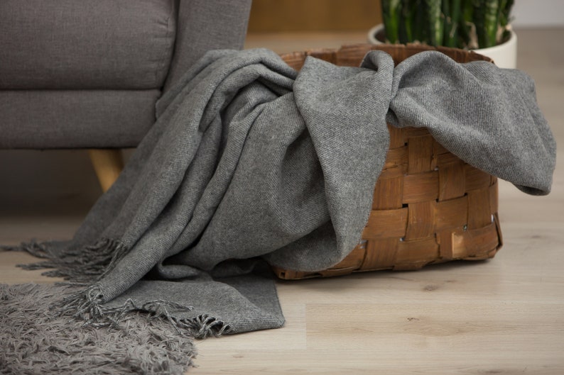 Puede incluir: Una manta de lana gris con flecos est&aacute; colocada sobre una cesta tejida. La cesta est&aacute; hecha de materiales naturales y tiene un color marr&oacute;n claro.