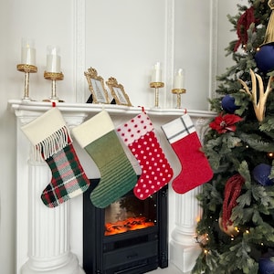 Peut inclure: Quatre chaussettes de Noël suspendues en rangée. La première est à carreaux verts, rouges et blancs avec un revers crème à franges. La deuxième est vert uni avec des rayures horizontales. La troisième est rouge à pois blancs. La quatrième est rouge uni.