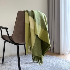 Könnte beinhalten: Eine grün-cremefarbene karierte Decke mit Fransen liegt über der Rückenlehne eines braunen Stuhls. Die Decke zeigt verschiedene Grün- und Cremetöne in einem Karomuster. Der Stuhl hat schwarze Beine und steht vor einem neutralen Hintergrund.