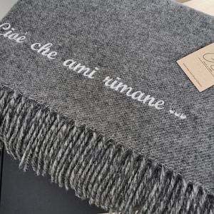 May include: A charcoal grey woven throw blanket featuring a fringed edge and the white printed text "Cioè che ami rimane..." across the surface. A rectangular tan paper tag with the logo "GS" is attached to the corner with a white string.