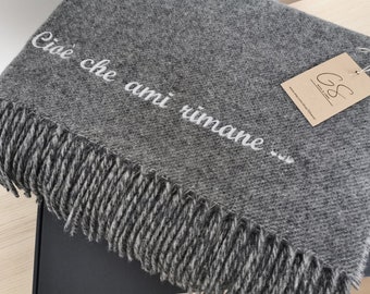 以下が含まれることがあります： 「Cioè che ami rimane...」という白い刺繍文字とフリンジの縁取りが施された、チャコールグレーの織りブランケット。ロゴ「GS」が印刷された長方形のベージュ色の紙タグが、白い紐で角に取り付けられています。