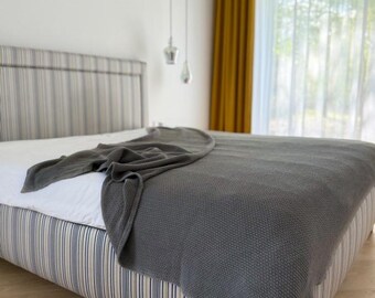 May include: A dark gray knitted throw blanket with a textured waffle pattern draped over a bed. The bed features a white sheet and a headboard and base upholstered in a fabric with vertical blue, yellow, and white stripes.