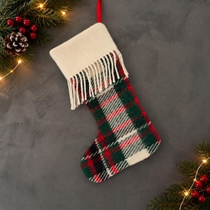 Peut inclure: Une chaussette de Noël avec un motif à carreaux dans des tons de rouge, vert foncé et blanc. Le revers supérieur est en tissu crème uni avec une bordure à franges. Une boucle en ruban rouge est fixée en haut pour suspendre la chaussette.