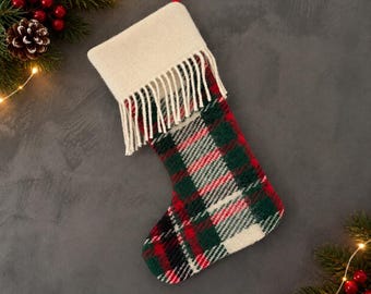May include: A Christmas stocking featuring a plaid pattern in shades of red, dark green, and white. The top cuff is solid cream-colored fabric with a fringe trim hanging down. A red ribbon loop is attached to the top for hanging the stocking.