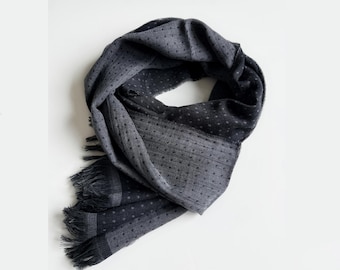 May include: A dark gray scarf featuring a subtle pattern of small, evenly spaced dots throughout the fabric. The scarf has a soft, textured appearance with frayed, fringed edges at the ends, creating a casual and lightweight look for the accessory.