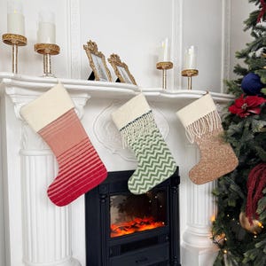 Op de afbeelding: Drie kerstsokken hangend aan een witte schoorsteenmantel. De linker sok is rood met horizontale strepen, de middelste is groen met een chevronpatroon en franjes, en de rechter is van gestructureerde beige stof met witte franjes.