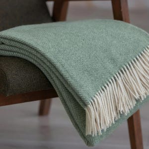 Puede incluir: Una suave manta de lana verde con un patrón de espiga y flecos blancos. La manta está colocada sobre una silla de madera.