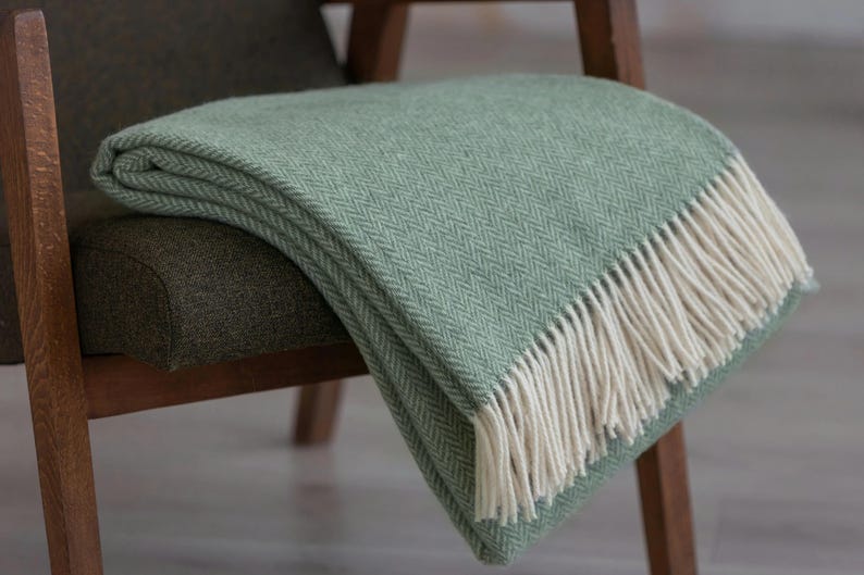Pu&ograve; includere: Una morbida coperta in lana verde con un motivo a spina di pesce e frange bianche. La coperta &egrave; drappeggiata su una sedia di legno.