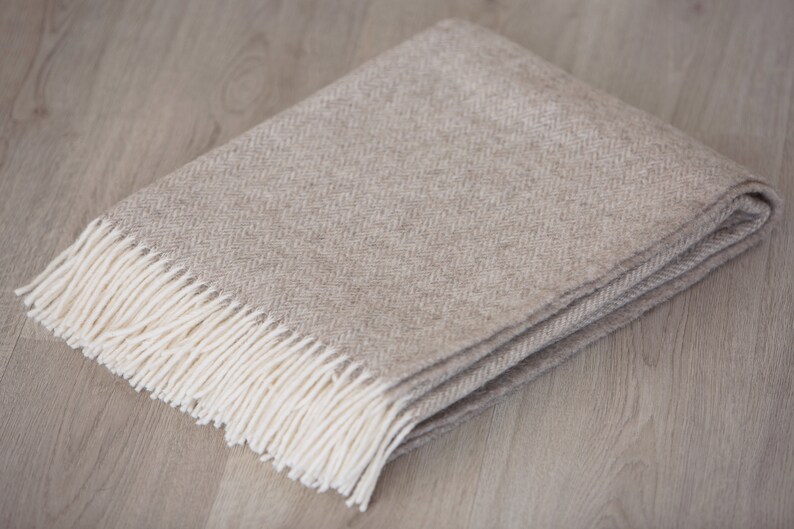 Pu&ograve; includere: Una coperta beige a spina di pesce con frange bianche.