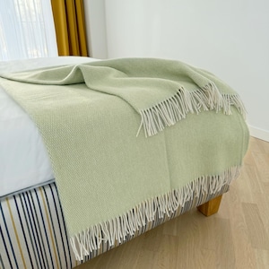 Könnte beinhalten: Eine hellgrüne gewebte Decke mit weißen Fransen ist über ein Bett drapiert. Die Decke hat ein strukturiertes Aussehen und besteht aus einem weichen Material. Das Bett hat einen gestreiften Bettrock.