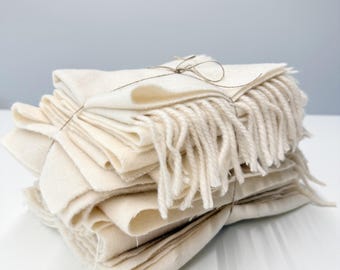 May include: A stack of folded off-white wool blankets tied together with thin brown twine. Each blanket features a soft, textured surface and a fringe edge consisting of multiple loose, hanging yarn strands along the side of the stack.