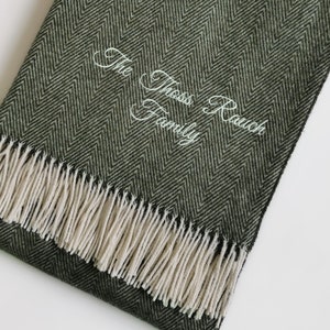 Könnte beinhalten: Eine dunkelgrüne, gewellte Wolldecke mit weißem Fransenbesatz und dem in Weiß gestickten Text "The Thass Rauch Family".