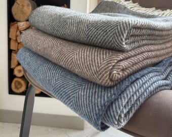 May include: A stack of four folded herringbone-patterned blankets in shades of gray, brown, and blue. The blankets are neatly arranged, with the top one showing a fringe. The blankets are on a chair.