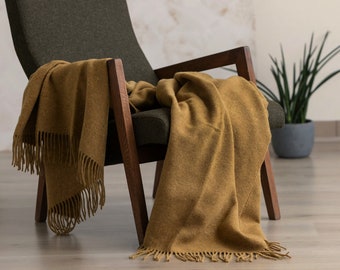 Puede incluir: Una manta de lana amarilla mostaza con flecos drapeada sobre un sillón verde oscuro. El sillón tiene brazos y patas de madera.