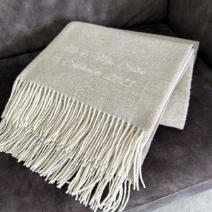 Peut inclure: Une couverture en laine gris clair avec une frange blanche. La couverture est brodée avec le texte "Mr. & Mrs. Saliba 15 septembre 2023".