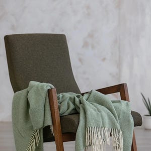 Pu&ograve; includere: Una coperta verde a spina di pesce con frange bianche drappeggiata su una poltrona verde scuro con braccioli in legno.