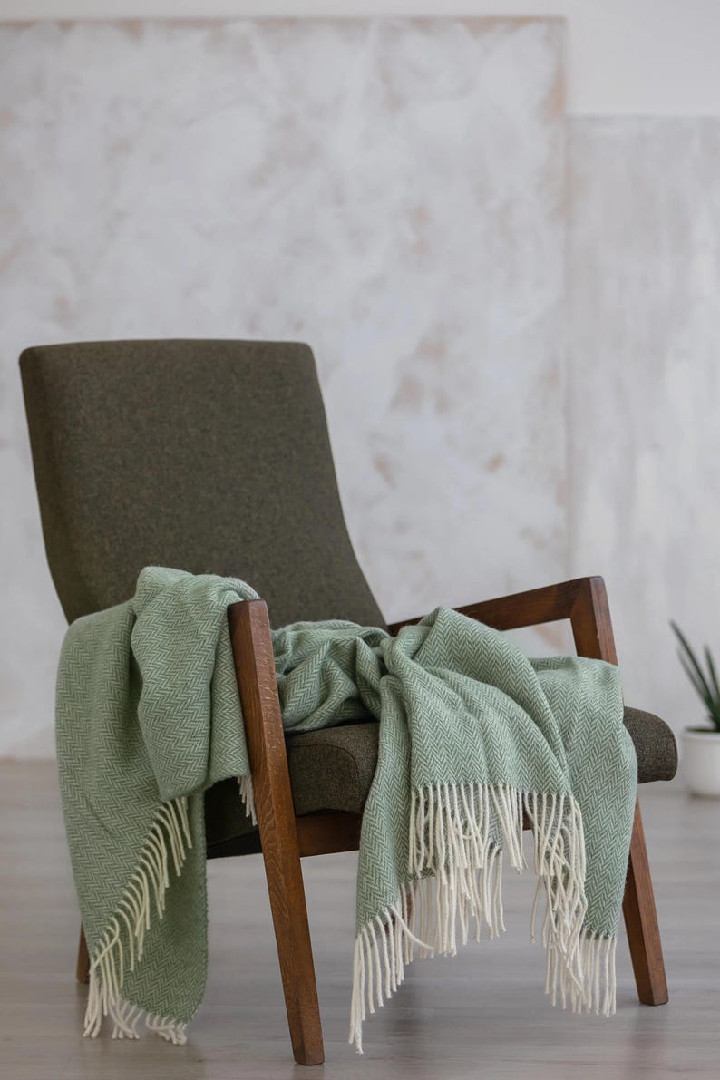 Pu&ograve; includere: Una coperta verde a spina di pesce con frange bianche drappeggiata su una poltrona verde scuro con braccioli in legno.