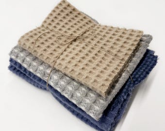 May include: A stack of three folded waffle-weave towels in beige, light gray, and dark blue. The towels are neatly layered and secured together with a thin, brown twine string tied in a simple bow across the center of the top beige towel.