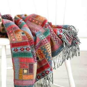 May include: A colourful woven throw blanket with fringe, draped over a white rocking chair. The blanket features a geometric pattern in shades of red, green, blue, and yellow.