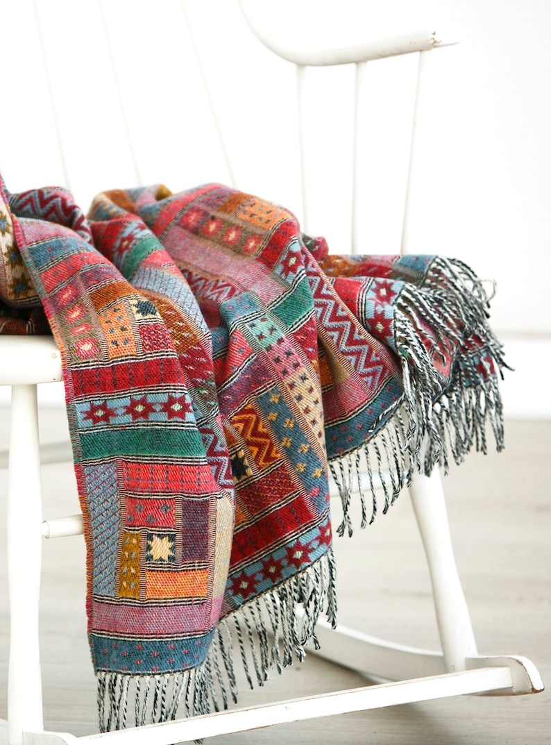 May include: A colourful woven throw blanket with fringe, draped over a white rocking chair. The blanket features a geometric pattern in shades of red, green, blue, and yellow.