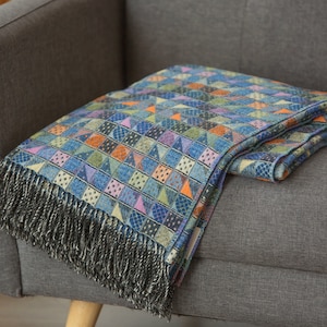 Könnte beinhalten: Eine blaue, grüne, orange und violette gemusterte Wolldecke mit schwarzen Fransen auf einem grauen Sofa.