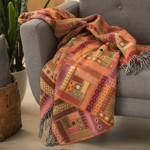 May include: A colourful throw blanket with a patchwork design in shades of orange, red, brown, and pink. The blanket is draped over a grey sofa.