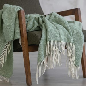 Pu&ograve; includere: Una morbida coperta verde chiaro con frange bianche, drappeggiata su una poltrona in legno. La coperta ha un motivo a spina di pesce.