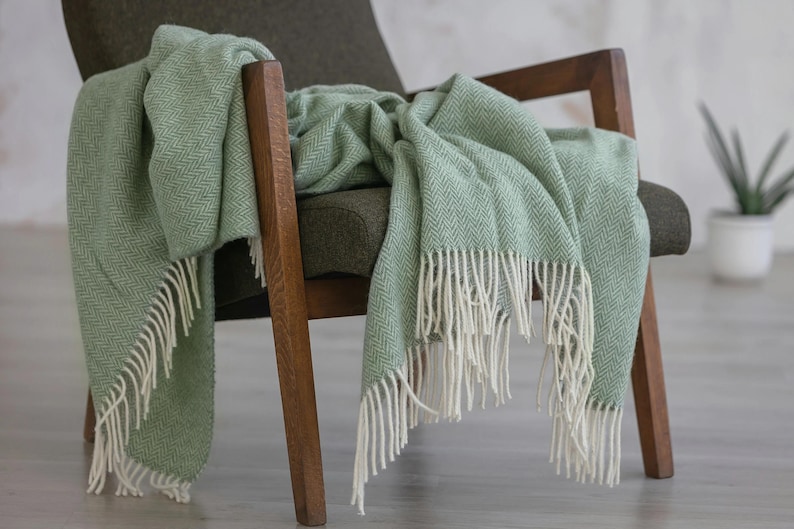 Pu&ograve; includere: Una morbida coperta verde chiaro con frange bianche, drappeggiata su una poltrona in legno. La coperta ha un motivo a spina di pesce.