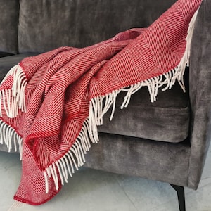 Peut inclure: Une couverture en laine rouge et blanche à motif chevron avec des franges blanches, posée sur un canapé gris.