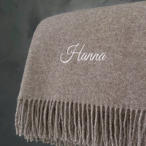 Peut inclure: Une couverture en laine de couleur marron avec une bordure en franges. La couverture est personnalisée avec le nom "Hanna" brodé en blanc.