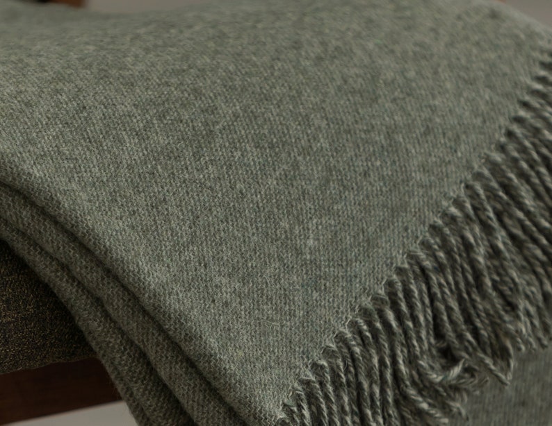 Puede incluir: Una suave manta de color verde con flecos. La manta est&aacute; doblada y colocada sobre una silla.