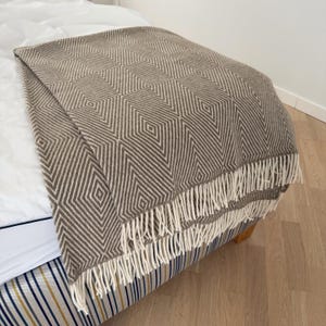 May include: A brown and white geometric patterned throw blanket with fringe. The blanket is folded over a bed, showcasing a diamond design. The fringe is a cream color. The blanket is made of a soft material.