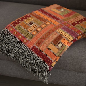May include: A colourful woven throw blanket with a geometric pattern in shades of orange, red, and brown. The blanket has a fringe border and is draped over a grey sofa.