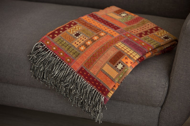 May include: A colourful woven throw blanket with a geometric pattern in shades of orange, red, and brown. The blanket has a fringe border and is draped over a grey sofa.