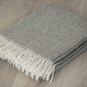 Könnte beinhalten: Eine graue und weiße Fischgrät-Wolldecke mit Fransenrand. Die Decke ist gefaltet und liegt auf einem Holzboden.