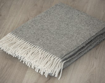 May include: A folded gray throw blanket featuring a woven herringbone pattern throughout the fabric. The blanket is finished with long, cream-colored fringe tassels along the edge, providing a textured contrast to the gray woven body.