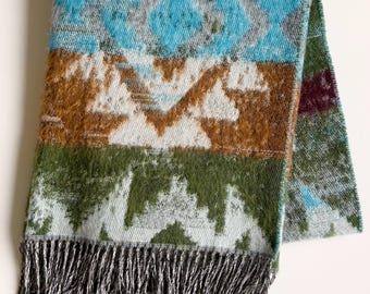 May include: A folded rectangular throw blanket featuring a horizontal color-blocked pattern in shades of bright turquoise, warm brown, and olive green. The fabric displays a repeating geometric diamond motif and includes a dark grey fringed edge.