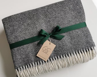 Puede incluir: Una manta de lana y lino gris y blanco con un patrón de espiga, con una cinta verde atada en un lazo. La manta tiene un borde de flecos y una etiqueta marrón con el texto "G&S Wool & Linen" y una dirección de sitio web.