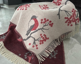 May include: A cream-colored throw blanket with a red bird and red berries pattern. The blanket has a burgundy underside and a fringe border.