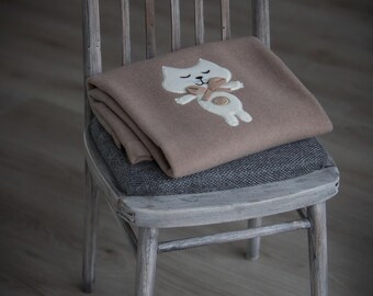 May include: A folded light brown blanket featuring a white cat applique with closed eyes, a small tan bow at the neck, and a circular tan detail on the belly. The blanket is draped over a wooden chair with a gray seat cushion.